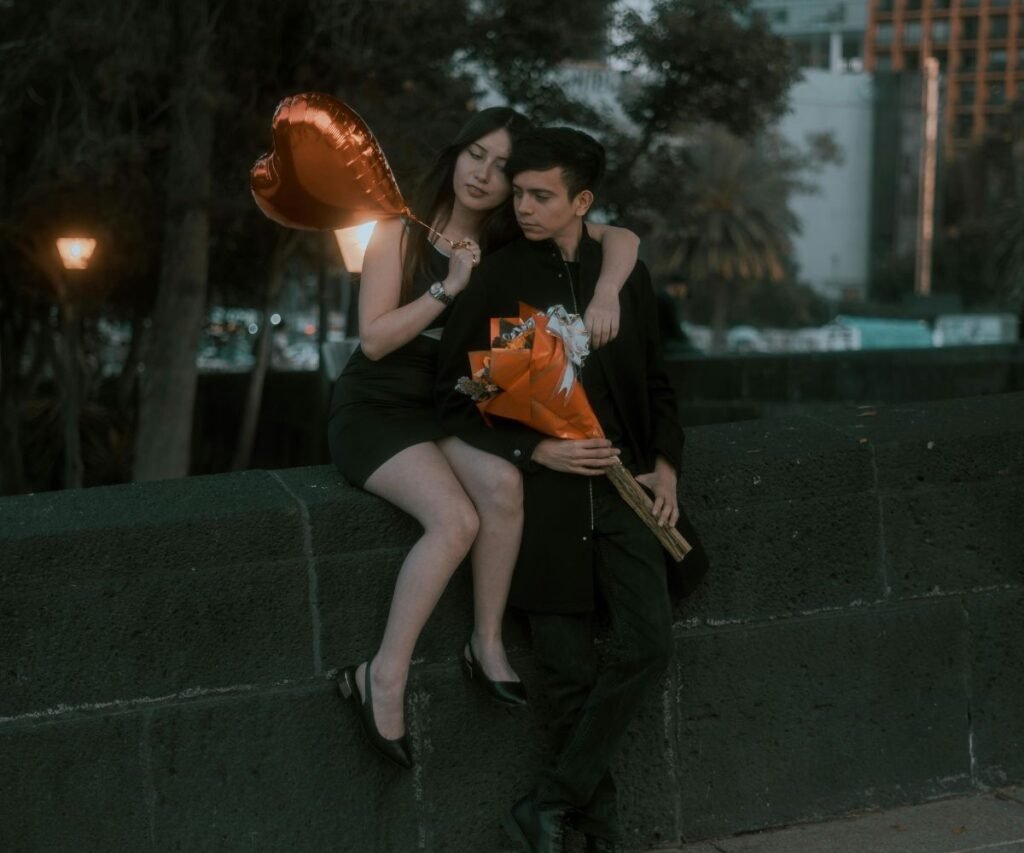 Romantic couple sitting together with flower bouquet and heart-shaped balloon at sunset in city park